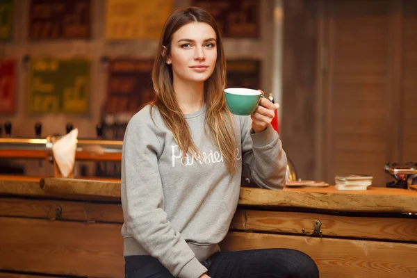 Fotó a szürke pulóver nő csésze kávé fából készült asztal közelében ült. — Stock Fotó