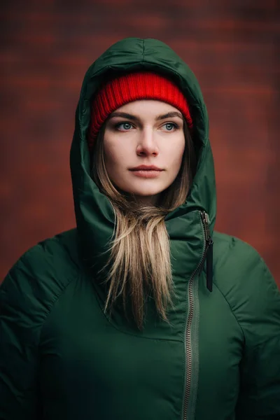Portrait of woman in green jacket and red hat on background of brick wall in afternoon. — Stock Photo, Image