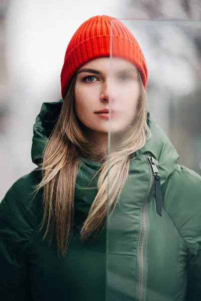 Портрет блондинки в зеленой куртке и красной шляпе возле стекла — стоковое фото