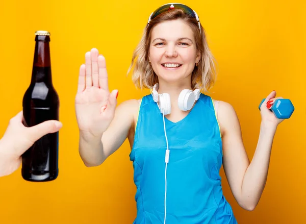 Chica atlética en auriculares con mancuerna en la mano delante de la mano con botella de cerveza . — Foto de Stock