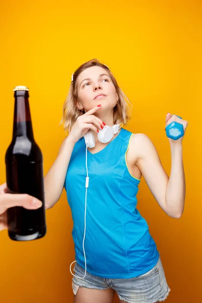 Chica atlética reflexiva en auriculares con mancuerna en la mano delante de la mano con botella de cerveza . — Foto de Stock