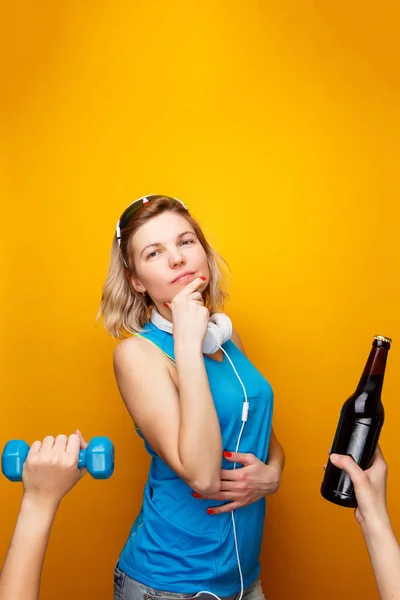 Задумчивая спортивная блондинка в наушниках перед выбором гантели или пивной бутылки . — стоковое фото