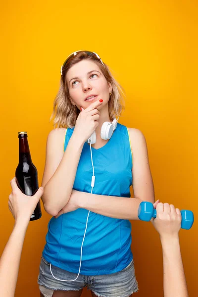 Задумчивая атлетичная девушка в наушниках перед тем, как выбрать гантель или бутылку пива — стоковое фото
