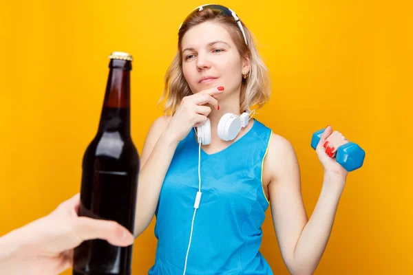Спортивная блондинка в наушниках с гантели в руке перед рукой с бутылкой пива . — стоковое фото