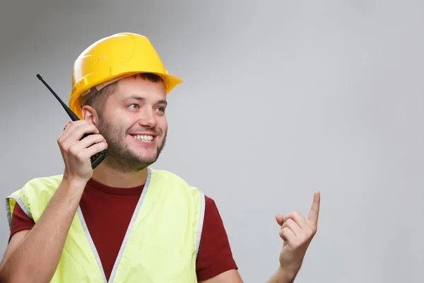 Foto de constructor alegre en casco amarillo hablando en walkie-talkie apuntando con el dedo hacia arriba — Foto de Stock