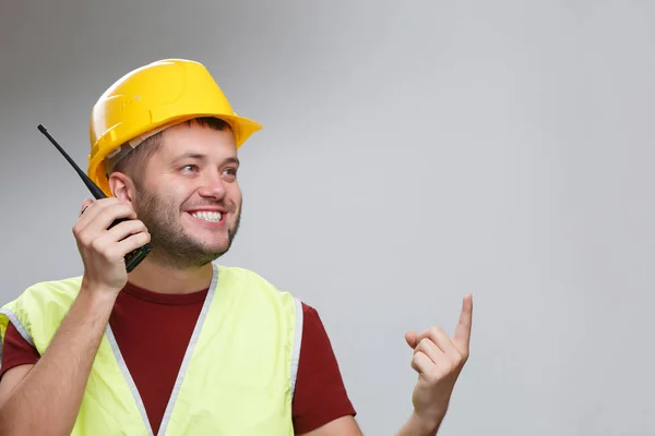 穿着黄色头盔的欢快建筑工人的形象在工作室里用对讲机指着他的手指. — 图库照片