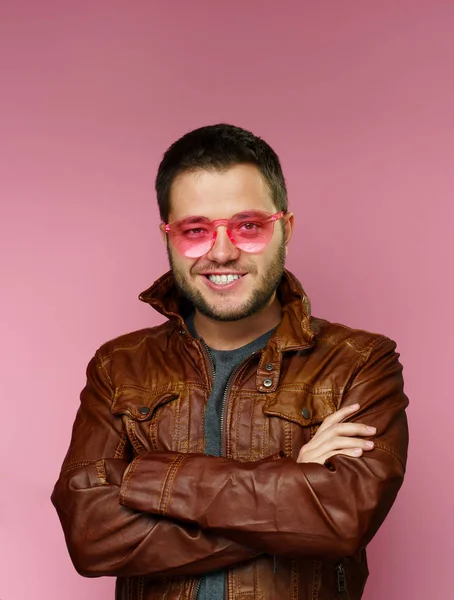 Zdjęcie uśmiechniętej Brunetki z różowymi okularami, skórzana kurtka, z ramionami skrzyżowanymi na różowym tle. — Zdjęcie stockowe