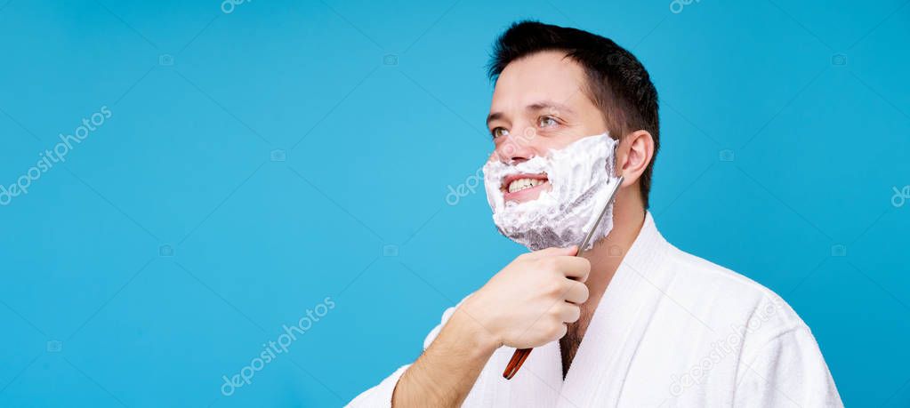 Photo of happy brunet man with straight razor in white coat on blank blue background in studio