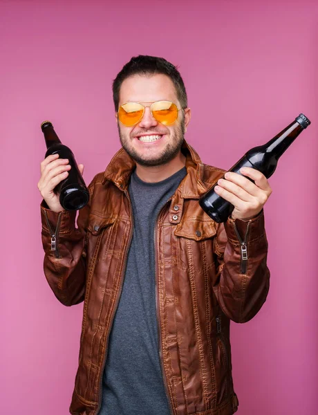 Foto de hombre sonriente en gafas naranjas y chaqueta de cuero con dos botellas en las manos sobre fondo rosa vacío — Foto de Stock