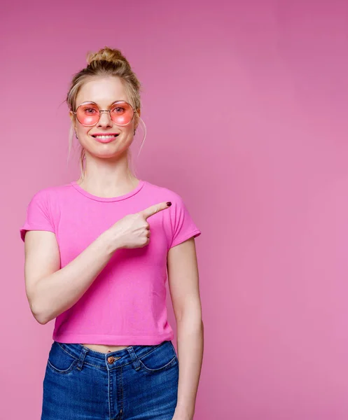 Fotografie usměvavé blondýny v růžových brýlích ukazujícími prstem na stranu — Stock fotografie