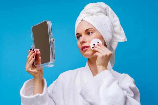 Afbeelding van jonge vrouw in witte vacht en handdoek op haar hoofd met wattenschijfje en spiegel in haar hand — Stockfoto