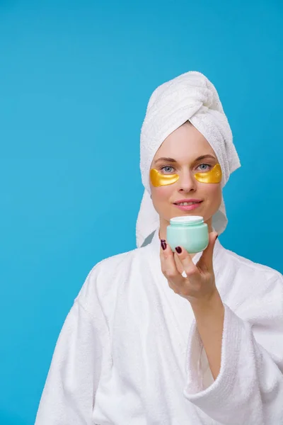 Изображение молодой женщины с гелевыми подушечками под глазами и полотенцем на голове и со сливками в руке — стоковое фото