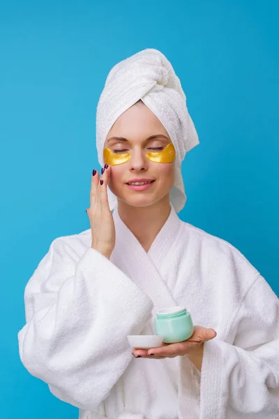Foto van jonge vrouw met gel pads onder de ogen en handdoek op hoofd en met crème in de hand — Stockfoto