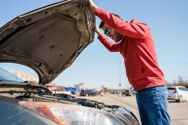 Image of side of man with glasses next to open hood of broken car during day