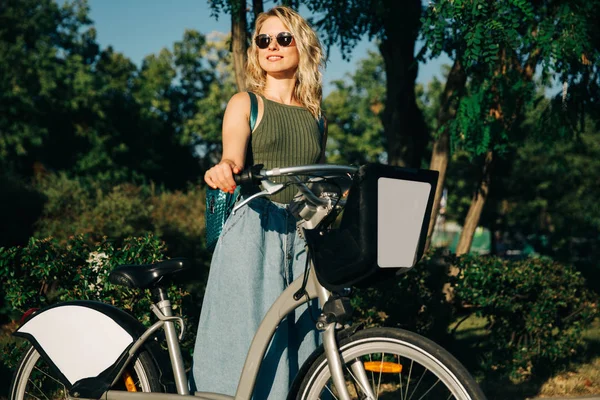 Fotó a boldog szőke napszemüveget nézett oldalon hosszú farmer szoknya állt kerékpárral mellett zöld bokrok a városban — Stock Fotó