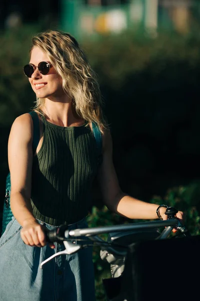 Imagen de la rubia rizada feliz en gafas de sol mirando al lado en falda de mezclilla larga de pie en bicicleta junto a arbustos verdes en la ciudad —  Fotos de Stock