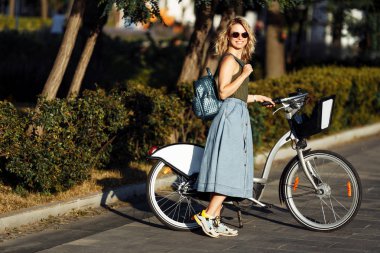 Yaz gününde şehirde yeşil çalılar yanında yolda bisiklet yanında ayakta uzun denim etek genç sarışın fotoğraf