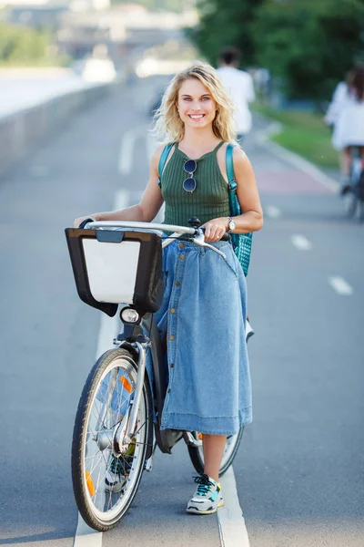 Fotó a fiatal szőke, hosszú denim szoknya mellett álló kerékpáros az úton a városban — Stock Fotó