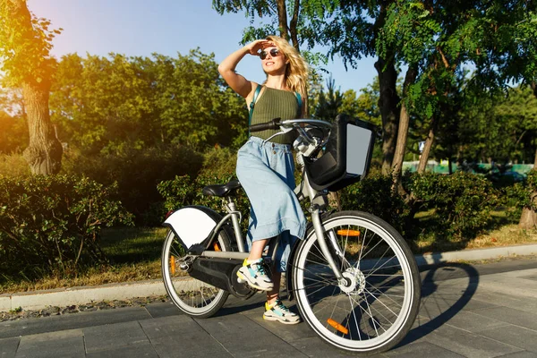 Pełna długość Zdjęcia Blondynka w okularach patrząc na bok w długiej spódnicy denim stoi na rowerze obok zielonych krzewów w mieście w letni dzień — Zdjęcie stockowe
