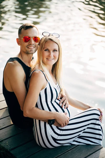 Nehir kıyısında oturan hamile sarışın kız ve erkek sarılma fotoğrafı — Stok fotoğraf