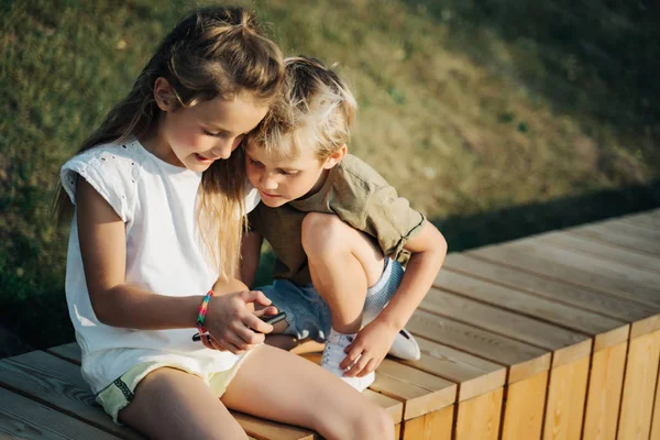 屋外の木製のフェンスに座って電話の手を持つ少年と女の子の写真 — ストック写真