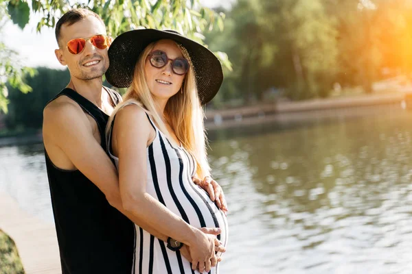 Foto van zwangere vrouw in hoed en mannen in roze bril staande op de oever van de rivier — Stockfoto