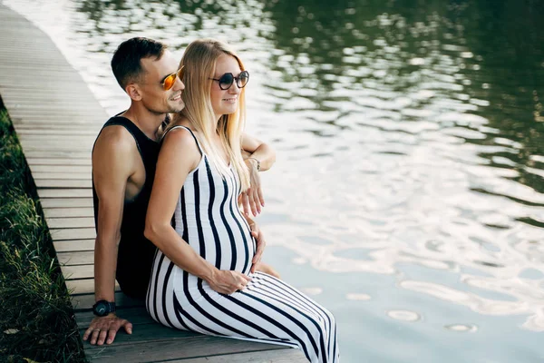 Foto van knuffelen zwangere blonde vrouw en mannen zittend op de oever van de rivier — Stockfoto