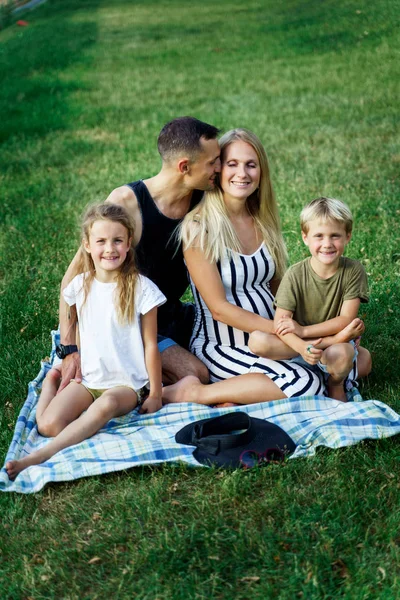 Picture of kissing parents with children sitting on plaid on picnic on summer day — Stock Photo, Image