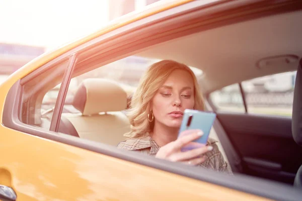 รูปถ่ายของสาวผมบลอนด์ที่มีโทรศัพท์ในมือของเธอนั่งในที่นั่งด้านหลังในรถแท็กซี่สีเหลืองในตอนบ่าย — ภาพถ่ายสต็อก