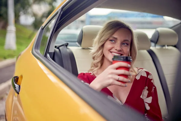 Foto de loira feliz com copo de café em suas mãos sentado no banco de trás do táxi amarelo — Fotografia de Stock