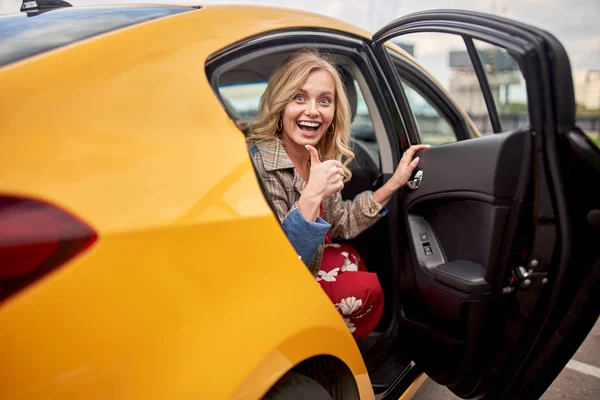 Foto de rubia entusiasta sentada en el asiento trasero de taxi amarillo con la puerta abierta —  Fotos de Stock