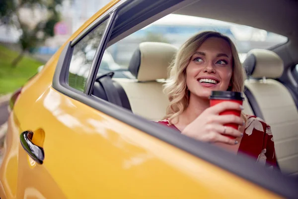 黄色いタクシーの後部座席に座っている彼女の手にコーヒーのグラスを持つ幸せなブロンドのイメージ. — ストック写真