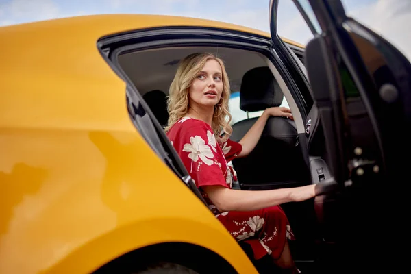 Foto de rubia en vestido rojo sentada en el asiento trasero de taxi amarillo con puerta abierta . —  Fotos de Stock