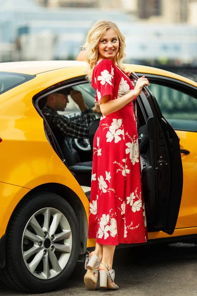Foto de larga duración de la mujer rubia en vestido rojo de pie cerca de taxi en verano  . —  Fotos de Stock