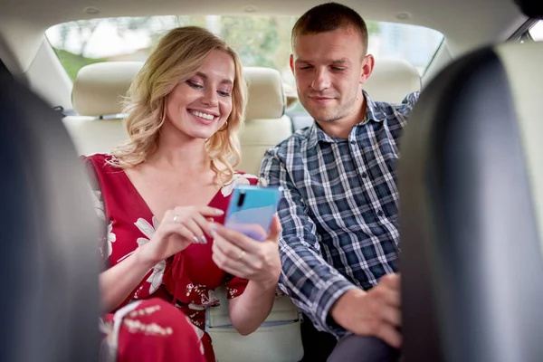 Foto de la mujer sonriente y el hombre con el teléfono en las manos sentado en el asiento trasero del coche —  Fotos de Stock