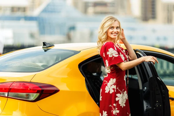 Изображение блондинки в красном платье, стоящей возле такси летом  . — стоковое фото