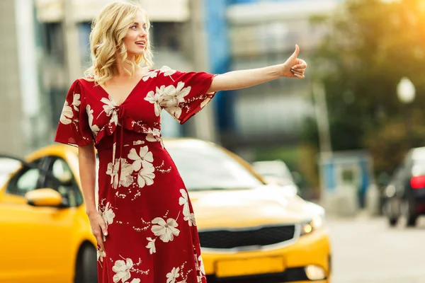Obraz blond w czerwonej sukni zatrzymując taksówkę na lato. — Zdjęcie stockowe