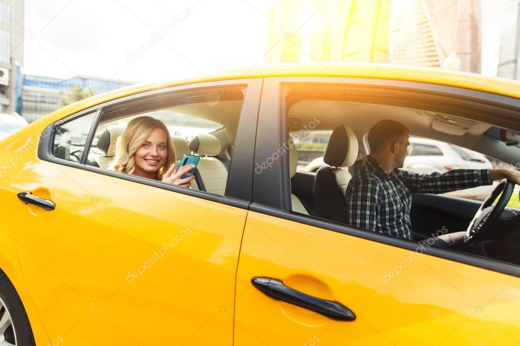 Photo of young blonde with phone in her hand sitting in back seat of yellow taxi with driver.