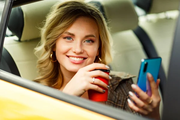 Photo of woman with coffee and phone in hands sitting in back seat of yellow taxi in summer on modern city background — Stock Photo, Image