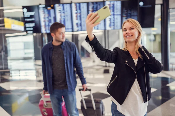 Image of man carrying luggage and woman taking selfie walking in waiting room at airport