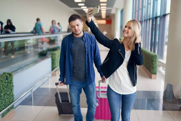 Foto del hombre llevando equipaje y la mujer tomando selfie caminando en la sala de espera en el aeropuerto — Foto de Stock