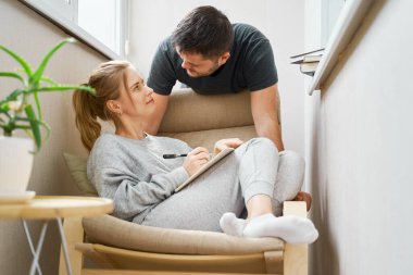 Sarışın, bej koltukta oturuyor ve balkonda yanında esmer duruyor.