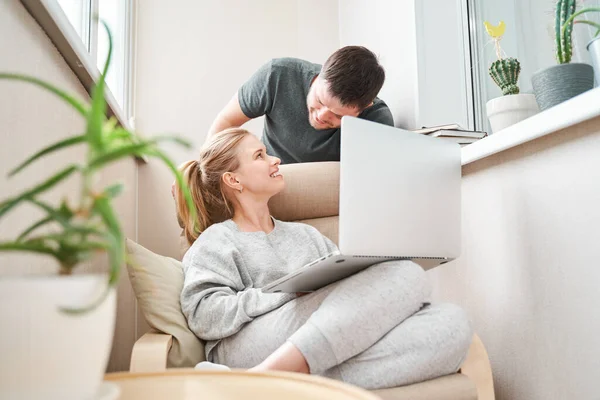 Щаслива жінка сидить на бежевому кріслі з ноутбуком і чоловіком в квартирі — стокове фото