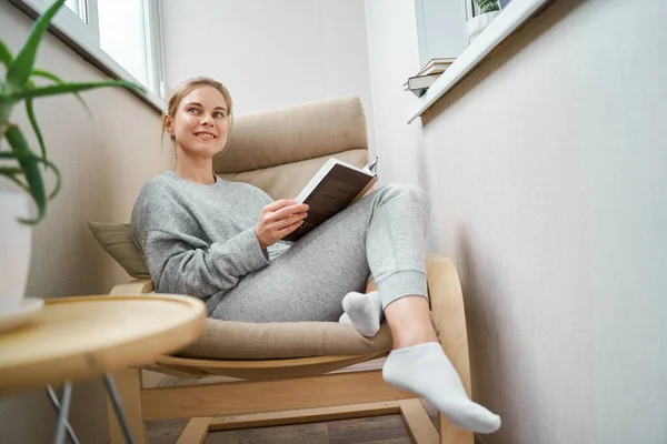 Menina feliz ler livro enquanto sentado em poltrona bege no apartamento — Fotografia de Stock