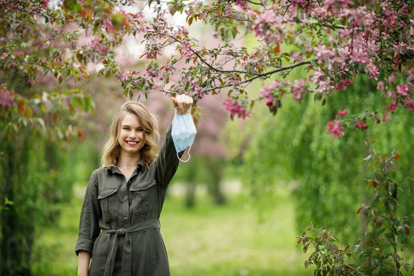 Mulher feliz em máscara médica no fundo de árvores floridas no parque à tarde . — Fotografia de Stock