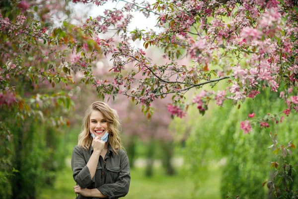 Mujer joven en máscara médica en el fondo de los árboles con flores en el parque por la tarde . — Foto de Stock