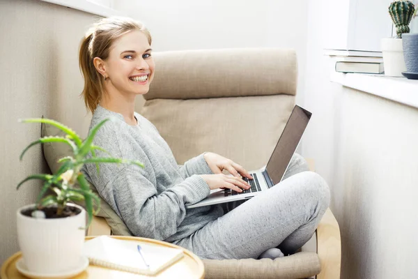 Щаслива блондинка в сірому домашньому костюмі з ноутбуком в руках, сидячи в кріслі . — стокове фото