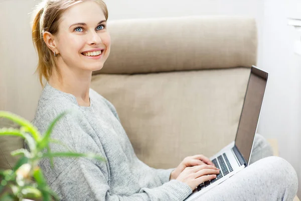 Щаслива блондинка в сірому домашньому костюмі з ноутбуком в руках, сидячи в кріслі — стокове фото
