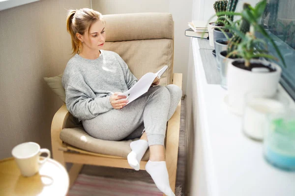 Fiatal nő olvasás könyvet, miközben ül a bézs karosszék a lakásban — Stock Fotó