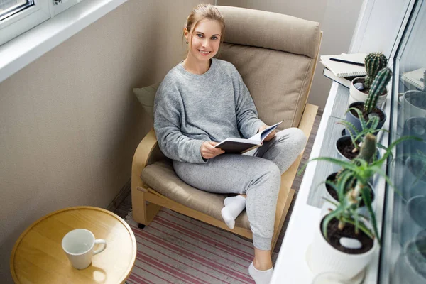Жінка з книгою в руках сидить на бежевому кріслі в квартирі — стокове фото
